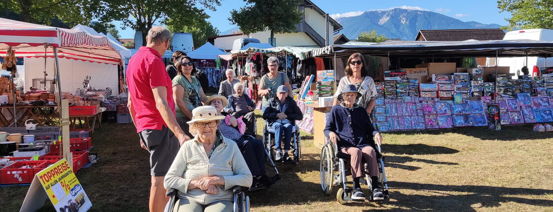 Senior*innen im Rollstuhl vor bunten Marktständen, Berge im Hintergrund
