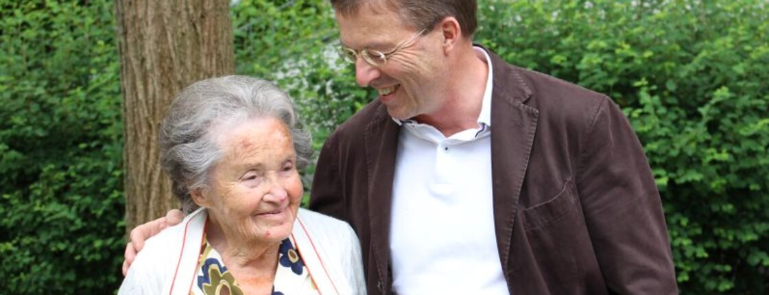 Ein Mann mit Brille steht neben seiner Mutter, hat den Arm um sie gelegt und sieht auf sie herab. Die beiden sind draußen vor einem Baum und Sträuchern.