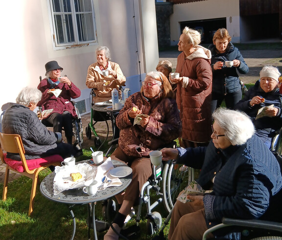 Ältere Menschen, einige in Rollstühlen, genießen eine Pause bei Tee oder Kaffee im Garten.