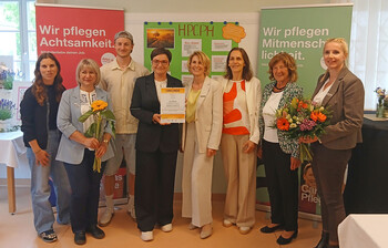 Gruppe aus Projektleitung, Pflege, Politik und Trägervertretungen steht mit Urkunde und Blumen vor HPCPH‑Tafel und Caritas‑Rollups