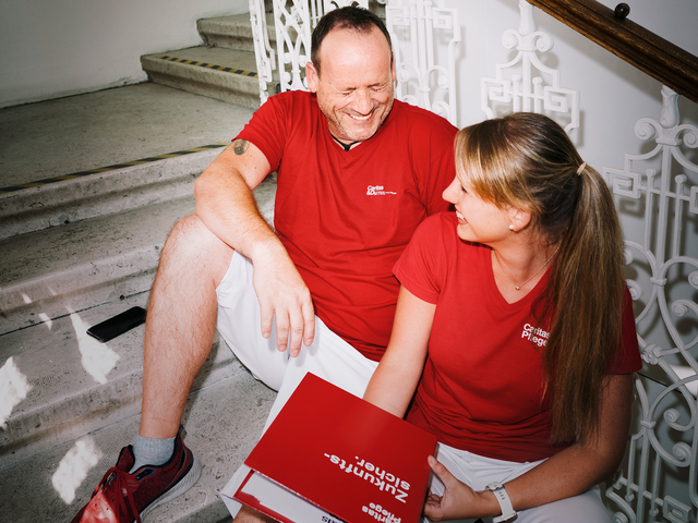 Ein Mann und eine Frau mit roten T-Shirts mit Caritas-Logo sitzen auf den Stiegen und lachen gemeinsam. Die Frau hat einen Aktenordner in der Hand.