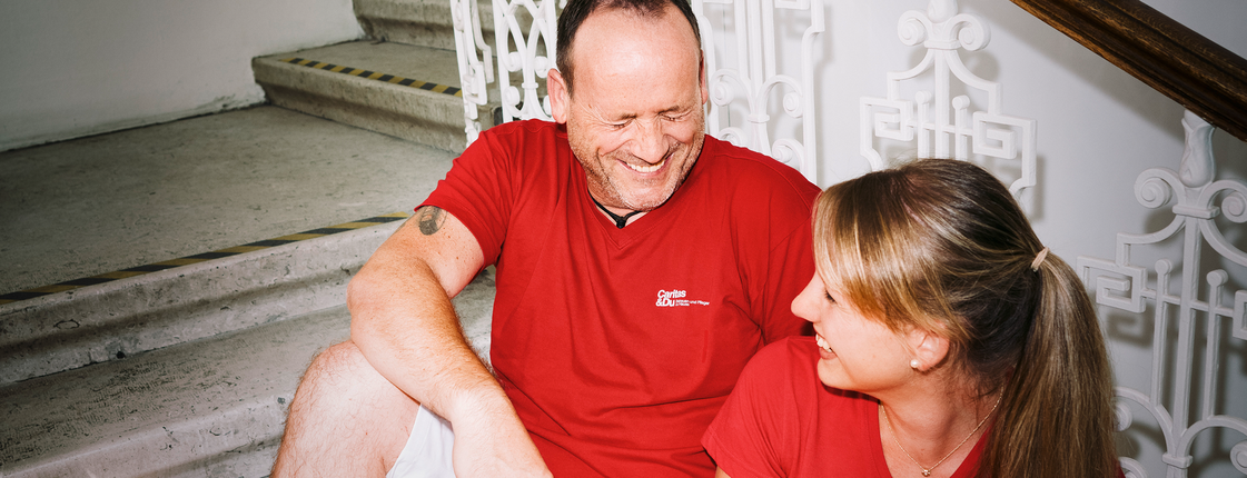 Ein Mann und eine Frau mit roten T-Shirts mit Caritas-Logo sitzen auf den Stiegen und lachen gemeinsam. Die Frau hat einen Aktenordner in der Hand.