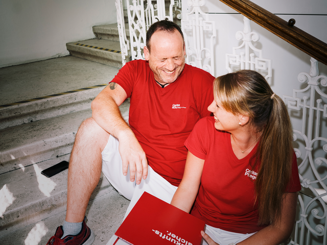 Ein Mann und eine Frau mit roten T-Shirts mit Caritas-Logo sitzen auf den Stiegen und lachen gemeinsam. Die Frau hat einen Aktenordner in der Hand.