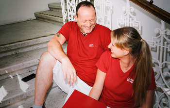 Ein Mann und eine Frau mit roten T-Shirts mit Caritas-Logo sitzen auf den Stiegen und lachen gemeinsam. Die Frau hat einen Aktenordner in der Hand.
