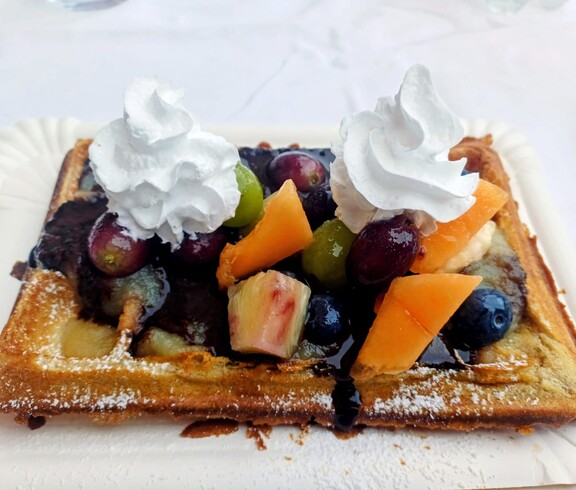 Eine frisch gebackene Waffel ist mit buntem Obst, Schokoladensoße und zwei Hauben Schlagobers garniert und auf einem weißen Teller angerichtet.