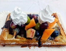 Eine frisch gebackene Waffel ist mit buntem Obst, Schokoladensoße und zwei Hauben Schlagobers garniert und auf einem weißen Teller angerichtet.