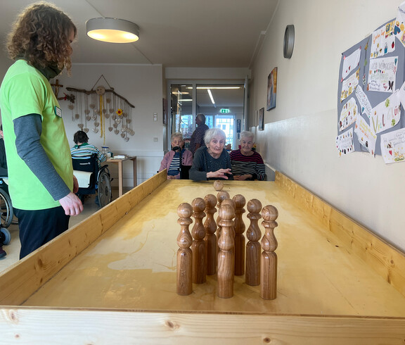 Bewohner*innen sitzen im Aufenthaltsraum und verfolgen das Tischkegeln auf der langen Holzbahn. 