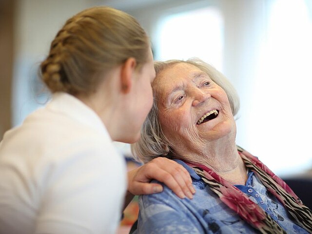 Eine junge Pflegerin beugt sich zu einer alten Dame, sie hat eine Hand auf deren Schulter gelegt. 