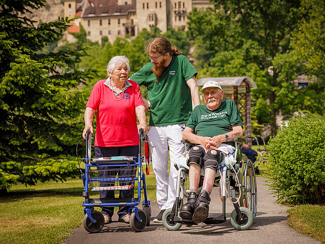Ein Pfleger steht mit einer Dame am Rollator und einem Mann im Rollstuhl im Garten des Hauses und lächelt die Dame an.