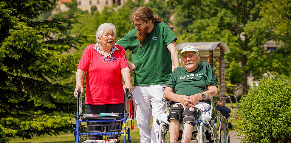 Ein Pfleger steht mit einer Dame am Rollator und einem Mann im Rollstuhl im Garten des Hauses und lächelt die Dame an.