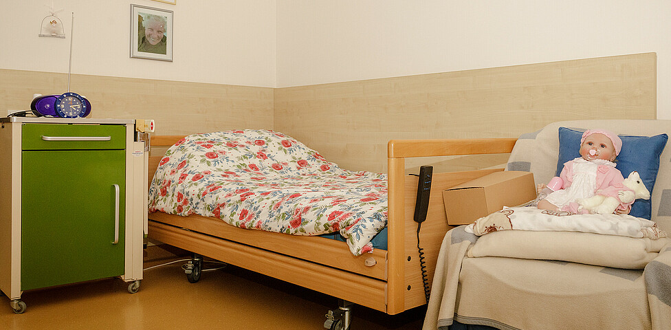 Ein Pflegebett, ein Nachtischt sowie ein bequemer Sessel auf dem eine Puppe sitzt, Bilder sind auf der Wand.