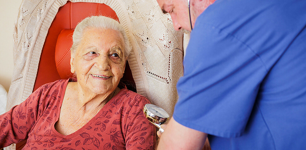 Ein Pfleger misst bei einer Dame den Blutdruck, während sie sich beide anlächeln.