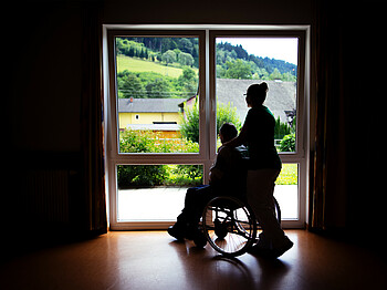 Eine Frau steht mit einem Menschen im Rollstuhl, die nur als Schatten gesehen werden, weil sie vor einem beleuchtenden Fenster stehen.