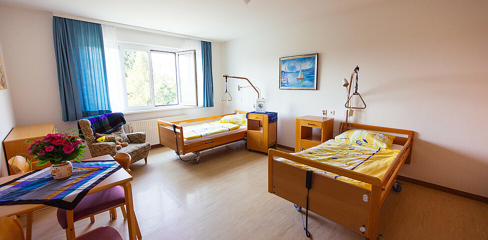 Ein Zimmer mit einem zwei Bett für pflegebedürftige Personen, 2 Nachtkästchen, 3 Sesseln, 1 Tisch mit Tischtuch und darauf stehender Blume sowie 1 Stuhl und 2 Bilder