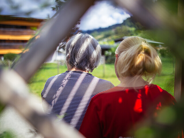 Eine ältere Dame und eine junge Frau sitzen nebeneinander in einer Laube, beide sieht man nur von hinten.