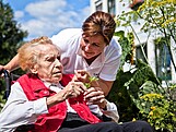Eine Caritas-Mitarbeiterin spricht mit einer Seniorin im Rollstuhl, die eine Blume in der Hand hält.