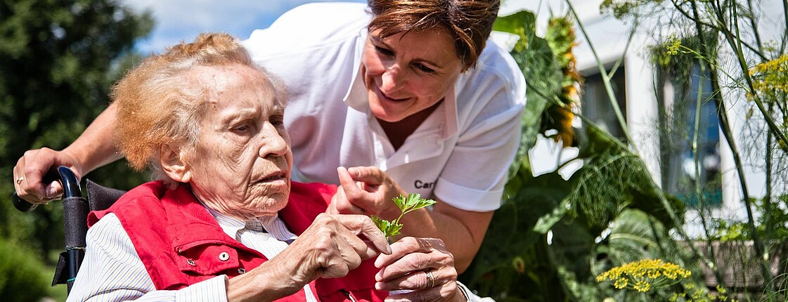 Eine Caritas-Mitarbeiterin spricht mit einer Seniorin im Rollstuhl, die eine Blume in der Hand hält.