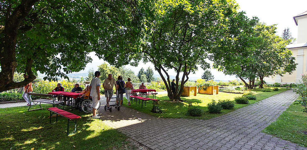 Auf der Wiese in der Gartenanlage befinden sich einige ältere Personen an 4 Tischen mit rotem Tischtuch. Schöne Bäume und 2 große Blumentröge befinden sich auch im Gartenbereich.