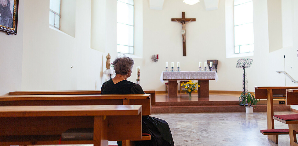 Eine ältere Dame sitzt in der hauseigenen Kapelle und blickt auf den Altar, auf dem sich 6 Kerzen und darunter ein Blumenstrauß befinden. Ein großes Christuskreuz hängt über dem Altar.