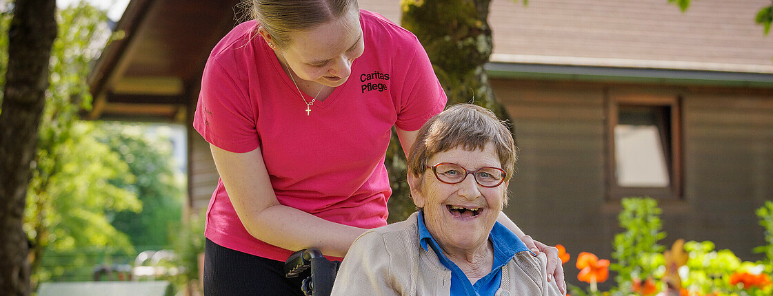 Eine Dame sitzt im Rollstuhl und lächelt, eine Pflegerin beugt sich über diese.
