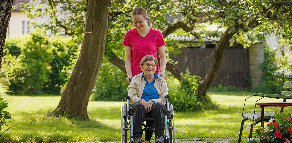 Eine Bewohnerin wird im Rollstuhl von einer Pflegerin durch den Garten geschoben.