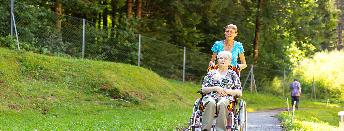 Eine Dame sitzt im Rollstuhl und eine junger Dame schiebt den Rollstuhl einen Weg entlang.