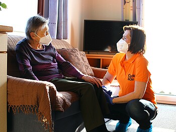 Mobile Pflegedienst, älter Frau links sitzt im Sessel, Pflegekraft rechts in der Hocke, sprechen miteinander