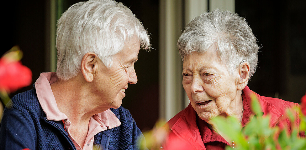 Zwei Bewohnerinnen stehen am Balkon und lachen gemeinsam. Vor ihnen kann man die Blumen des Gartens erkennen.