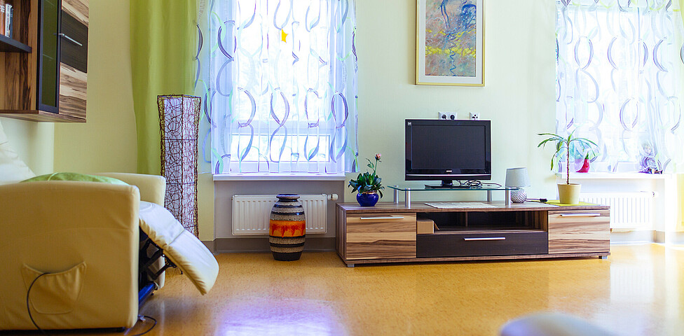 Ein Fernseher steht  auf einem Fernsehtisch. 2 Zimmerpflanzen befinden sich auch darauf. Am Boden steht eine Vase, daneben steht eine große Lampe. Ein Stuhl befindet sich unter einem Wandschrank. Über dem Fernseher hängt ein Bild. Links und rechts vom Fernseher befindet sich ein Fenster. Beide Vorhänge sind zugezogen.

