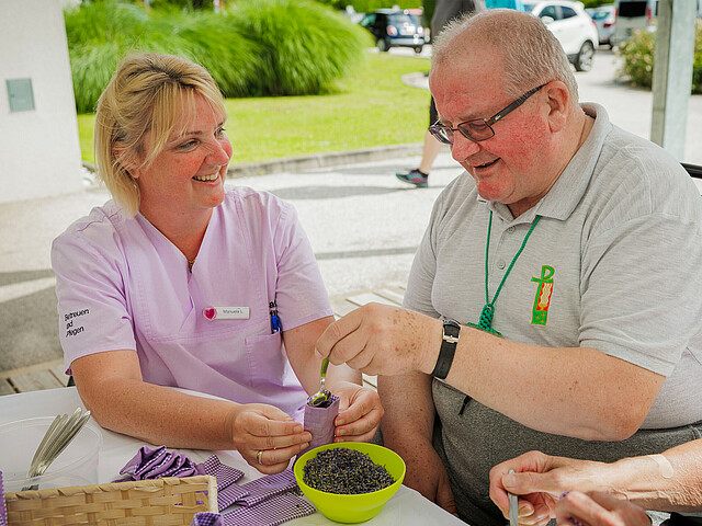 Eine Pflegerin sitzt bei einem älteren Mann und hilft ihm dabei Lavendelblüten in einen Beutel zu füllen.
