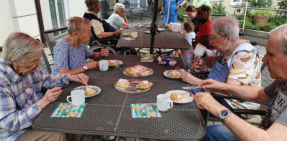 Gruppe älterer Leute sitzt beim Tisch im Garten bei Kaffee und Kuchen.