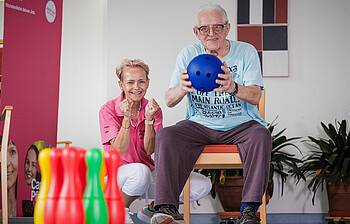 Ein Mann hält eine weiche Bowling-Kugel in den Händen, damit er die Pins im Vordergrund treffen kann. Eine Pflegerin kniet neben ihm und feuert ihn an.