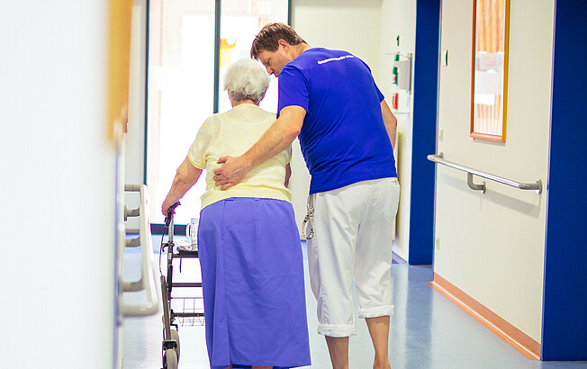 Ein Pfleger stützt eine ältere Dame beim Gehen mit ihrem Rollator.