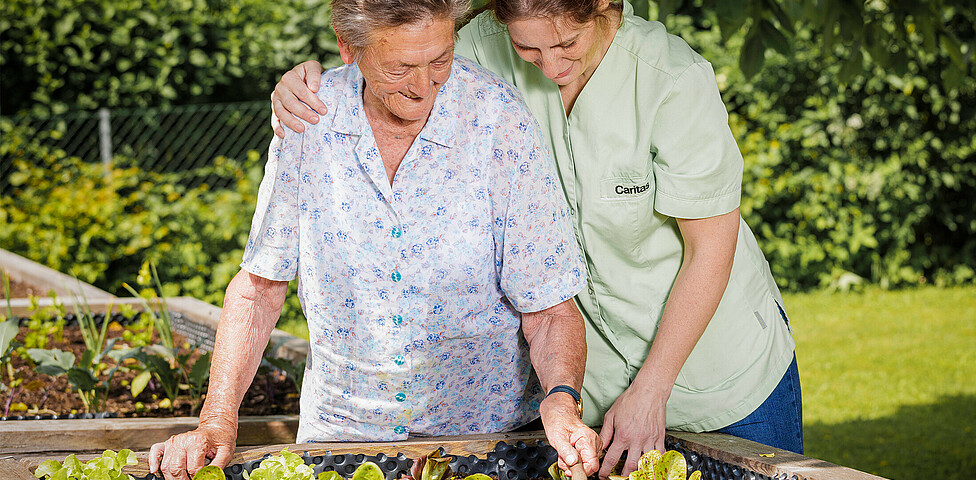Eine Pflegerin steht mit einer Bewohnerin an den hauseigenen Hochbeeten und kümmert sich um die Salatpflanzen.