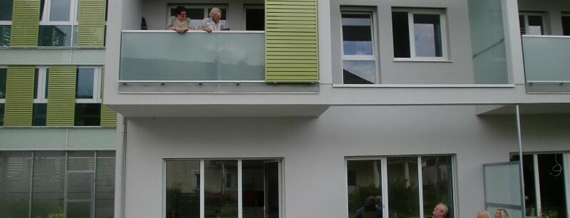 Ansicht des Hauses, auf einem Balkon im ersten Stock stehen zwei Frauen uns sehen auf zwei Frauen uns einen Mann herunter, die vor dem Haus auf einer Bank sitzen. 