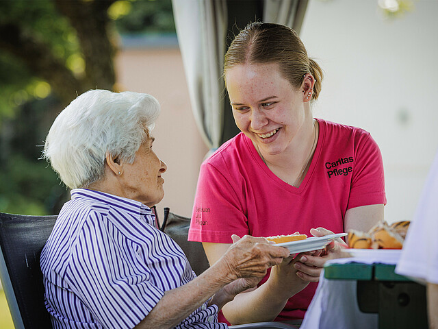 Eine Pflegerin reicht einer Dame einen Teller mit Kuchen, während sie sich gegenseitig anlächeln.