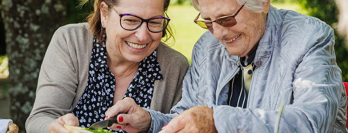 Zwei ältere Damen sitzen im Garten und schneiden Kräuter, beide lächeln.