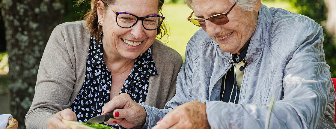 Zwei ältere Damen sitzen im Garten und schneiden Kräuter, beide lächeln.