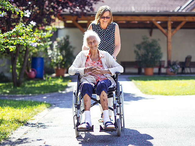 Eine junge Frau schiebt eine ältere Dame im Rollstuhl auf einem Weg im Garten entlang.