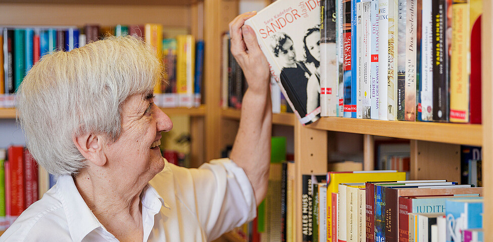 Eine Dame lächelt und zieht ein Buch aus einem Bücherregal.