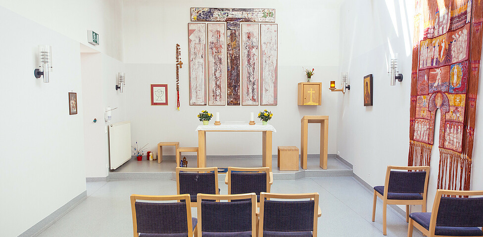 In diesem Raum befindet sich ein Altar bestehend aus einem Tisch mit 2 Kerzen und 2 Blumen. Bilder hängen an der Wand. Mehrere Stühle befinden sich in diesem Raum.