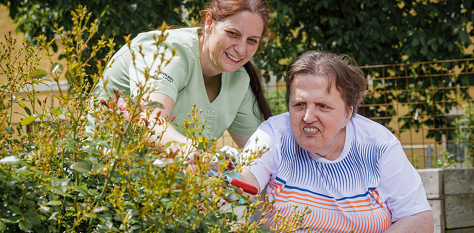 Eine Pflegerin hilft einer Bewohnerin dabei die Rosenknospen im Garten mit einer Gartenschere zu stutzen.
