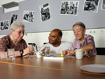 Zwei Seniorinnen sitzen am Tisch und malen, Pfleger in der Mitte