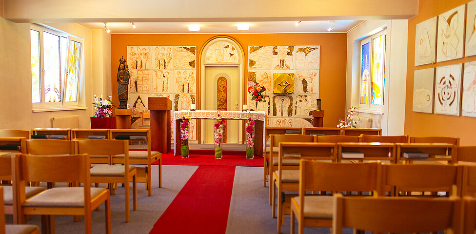 Ein langer roter Teppich führt zu einer schönen Kapelle. Es befindet sich ein Altar mit Kerzen, Blumen, Figur der Heiligen Maria mit Jesuskind, Gemälden, bunten Kirchenfenstern und ausreichend Sitzgelegenheiten.