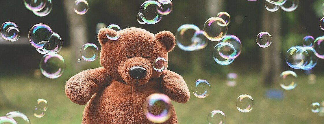 Brauner Teddybär sitzt mit geschlossenen Augen in Mitten von Seifenblasen.