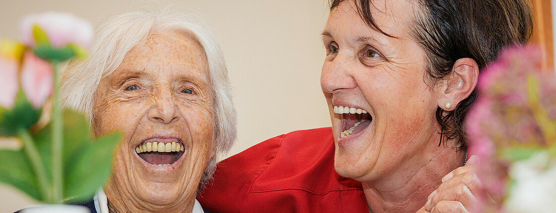 Eine ältere Dame lacht herzlich mit einer Pflegerin, dabei halten sie vertraut Hände
