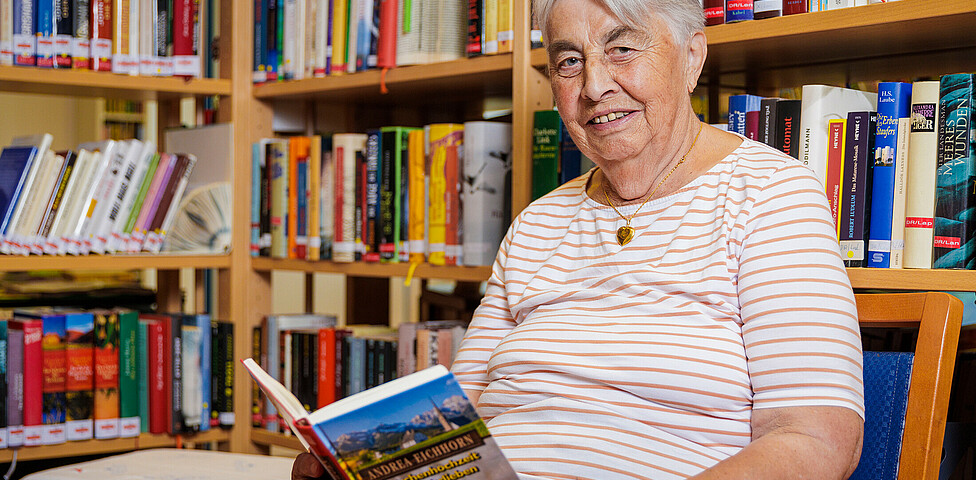 Eine ältere Dame sitzt vor einem Bücherregal, hat ein aufgeschlagenes Buch in der Hand und lächelt in die Kamera.