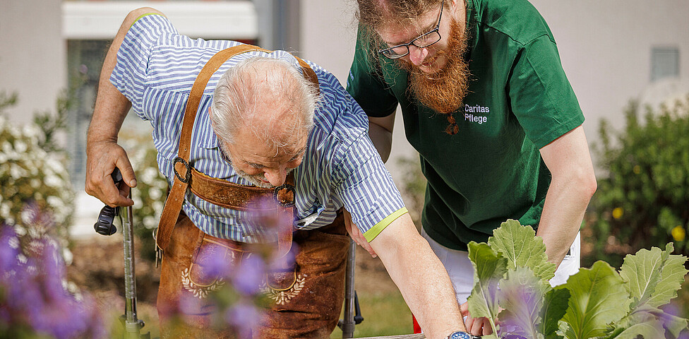 Ein älterer Mann arbeitet mit der Hilfe eines Pflegers in einem Hochbeet des Gartens.