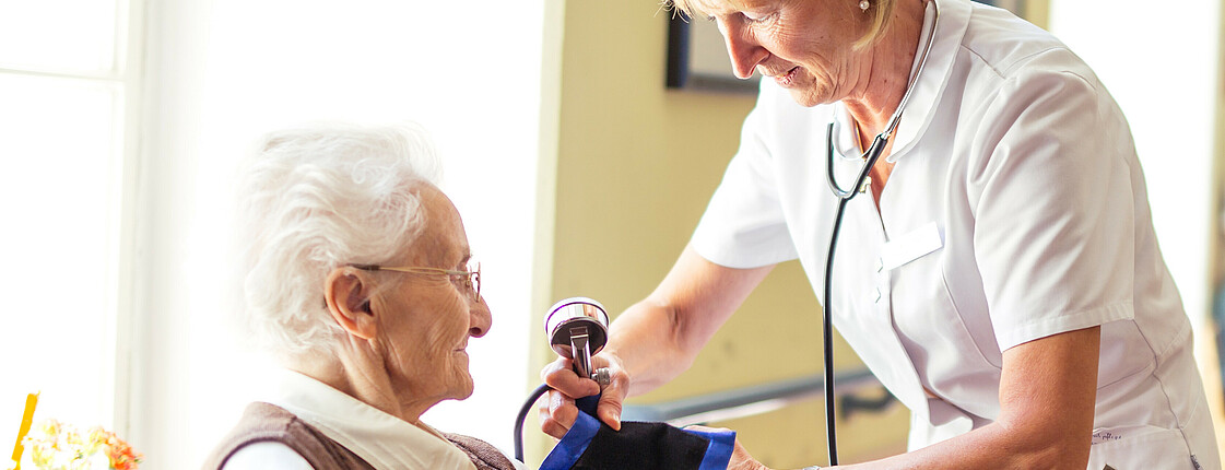 Eine Diplomierte Gesundheits- und Kranenpflegerin misst bei einer älteren Dame im Rollstuhl den Blutdruck.