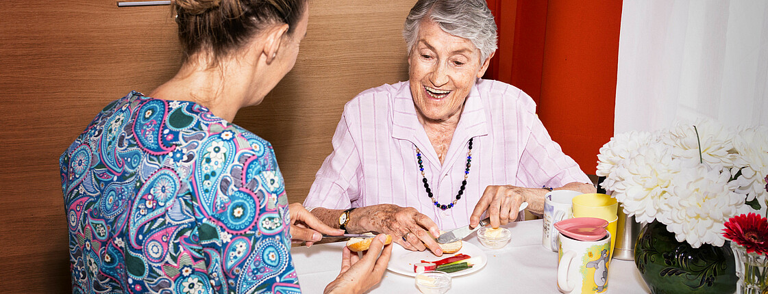Eine Caritas Mitarbeiterin und eine ältere Dame sitzen gemeinsam an einem Tisch und unterhalten sich, die ältere Dame lacht.